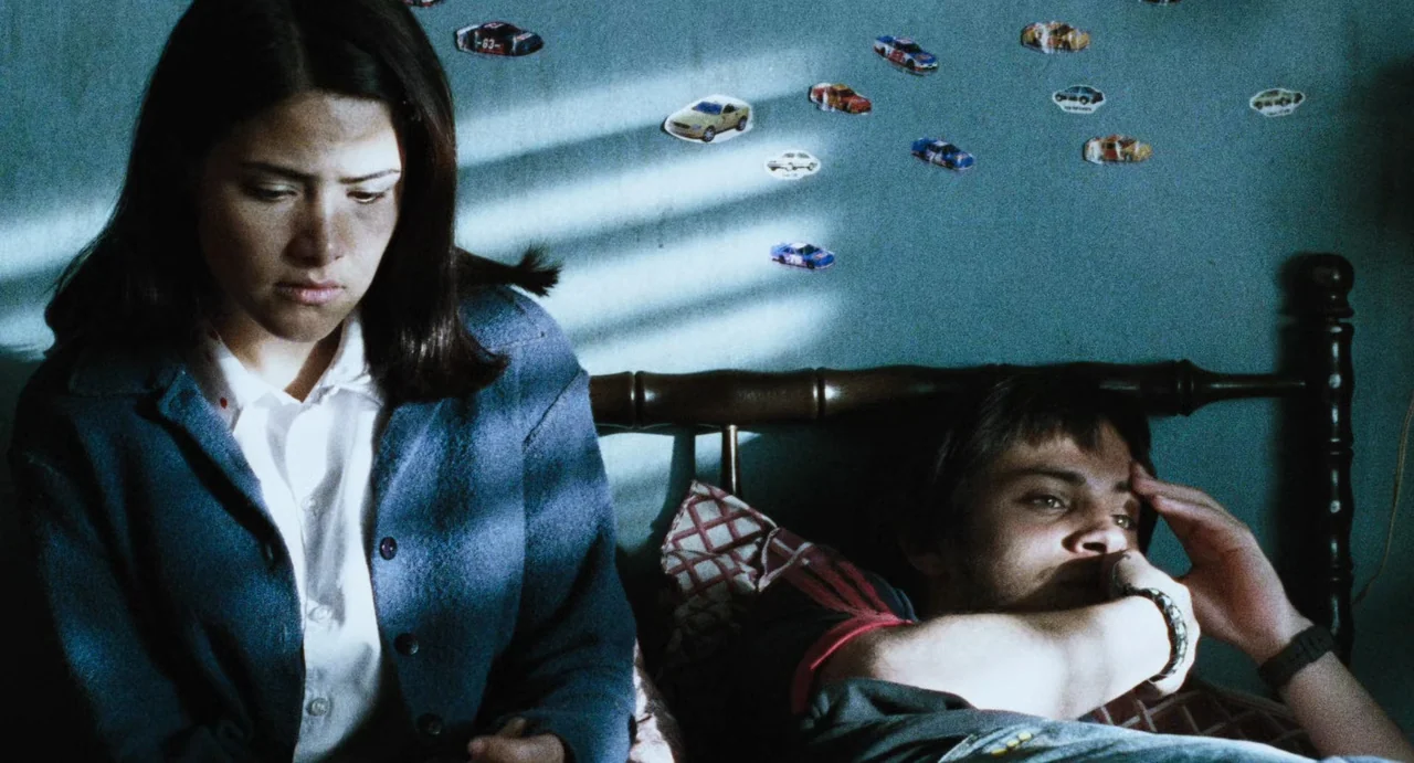 Amores Perros (2000) movie still: medium — A dimly lit room with walls decorated with small stickers of race cars. A woman sits on the edge of the…