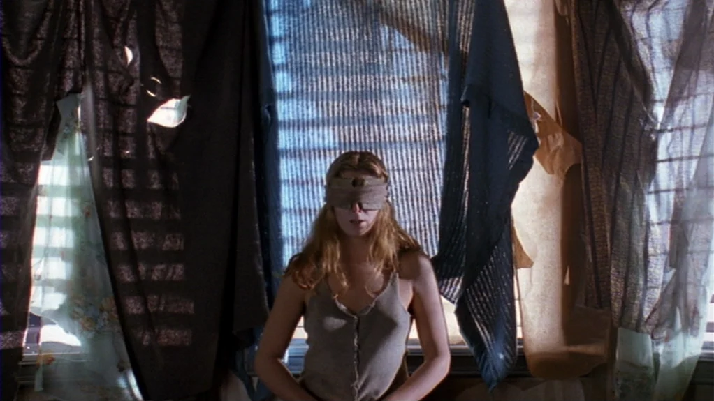 Patty Hearst (1988) movie still: medium — A woman stands before a window covered by multiple layers of hanging, worn fabric. She is blindfolded and…