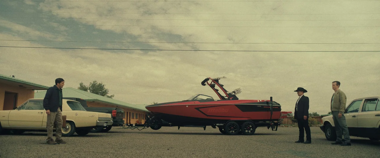 LaRoy, Texas (2023) movie still: wide — A gravel motel parking lot under an overcast sky features a red speedboat trailer. Three men stand near two…