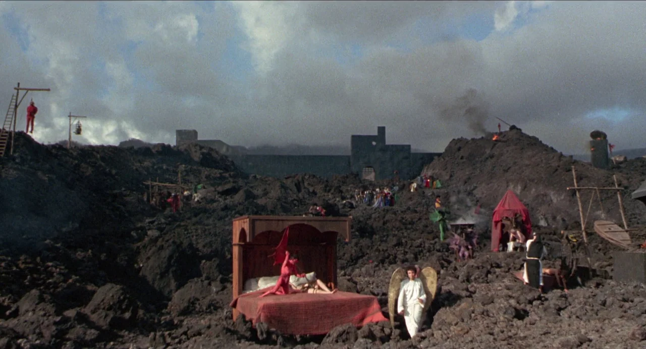 The Canterbury Tales (1972) movie still: wide — A vast volcanic landscape with dark jagged lava rock and a distant fortress. A wooden bedframe sits…