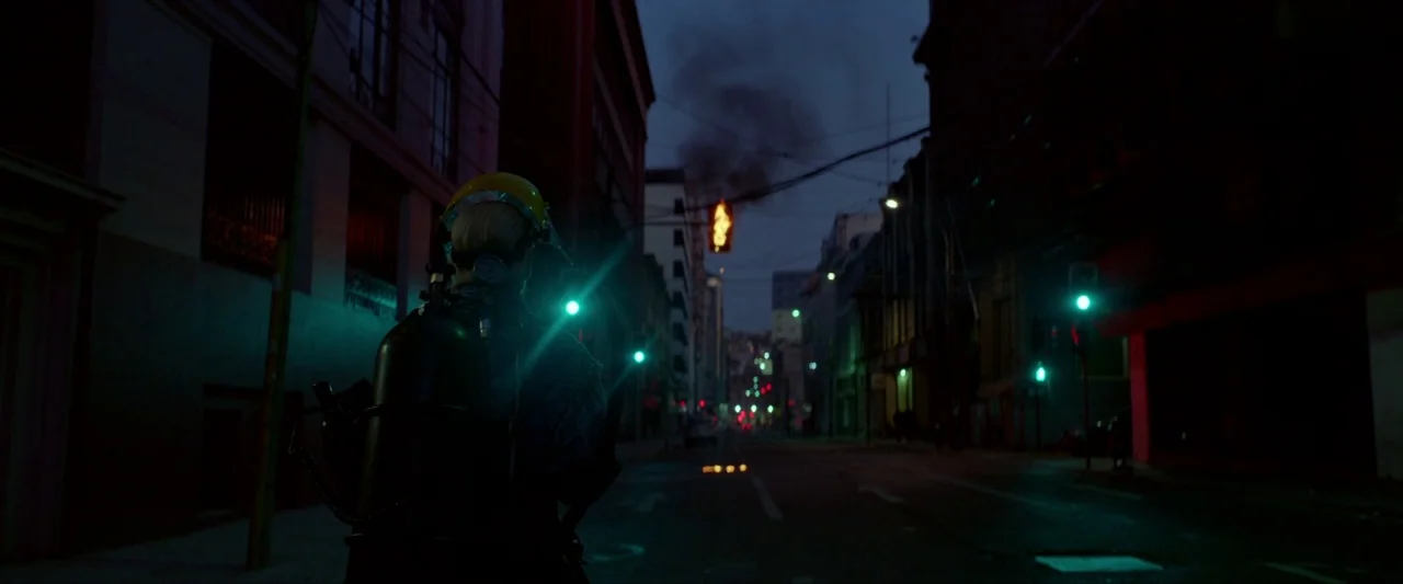 Ema (2019) movie still: medium — A deserted urban intersection at night features dark buildings flanking a central road. A person wearing protective…