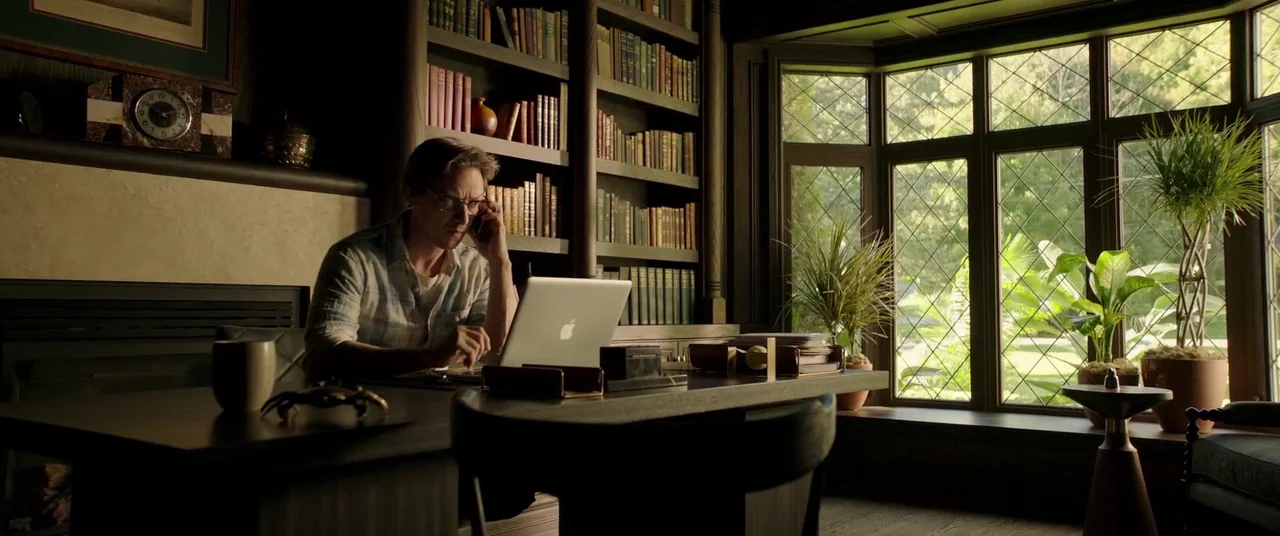 It Chapter Two (2019) movie still: medium — A dark wood panelled room filled with bookshelves surrounds a large desk with a laptop. A man sits at…