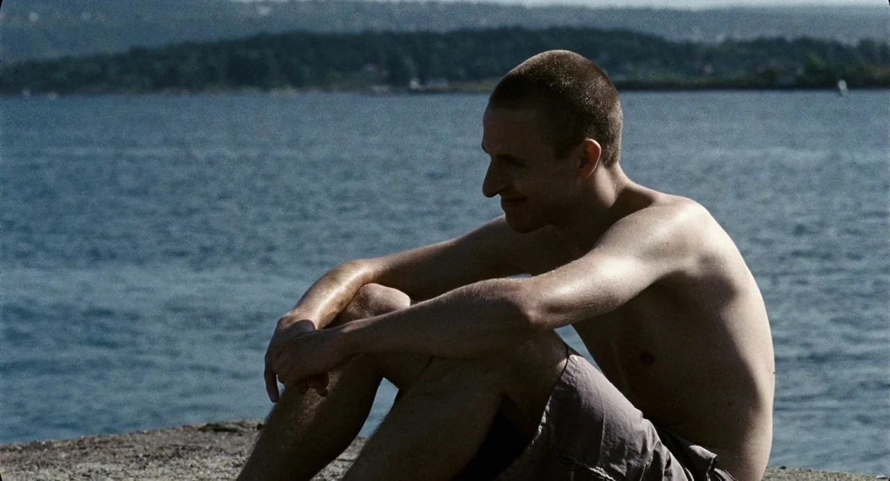 Reprise (2006) movie still: medium — A man sits on a concrete edge with a large body of water behind him. The man is wearing swim trunks and looks…