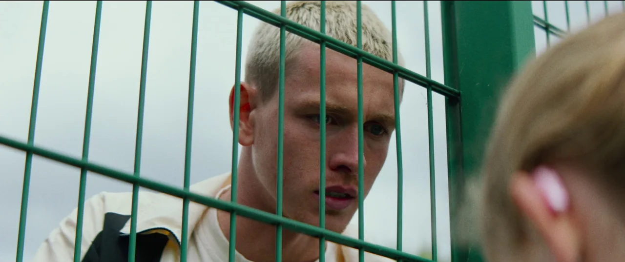 Scrapper (2023) movie still: closeup — A man looks through a green metal wire mesh fence at another person. The background is slightly blurred and…