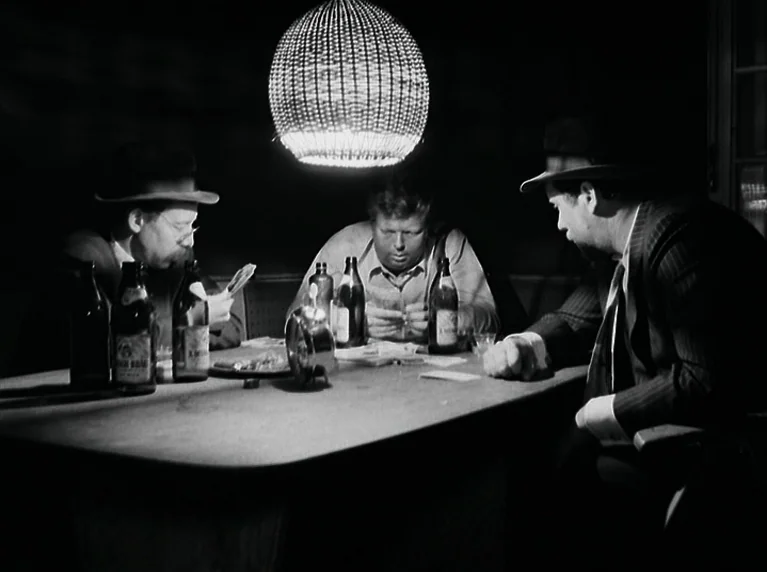 The American Soldier (1970) movie still: medium — A dimly lit interior with three men huddled around a table playing cards. A single woven lamp…