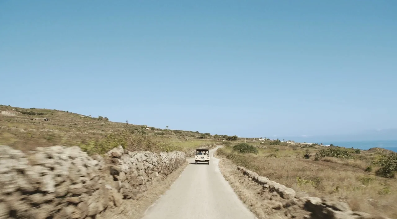 A Bigger Splash (2015) movie still: wide — A rural landscape features a paved road bordered by dry stone walls. A small open-top vehicle drives…