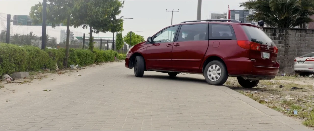 Saving Onome (2024) movie still: wide — A red minivan sits parked on a paved roadside next to a brush line. The vehicle is angled toward the edge of…
