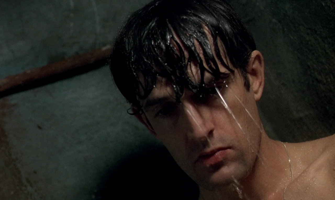 Dellamorte Dellamore (1994) movie still: closeup — A dimly lit tiled shower stall with dark, textured wall surfaces. A man with wet dark hair stands…