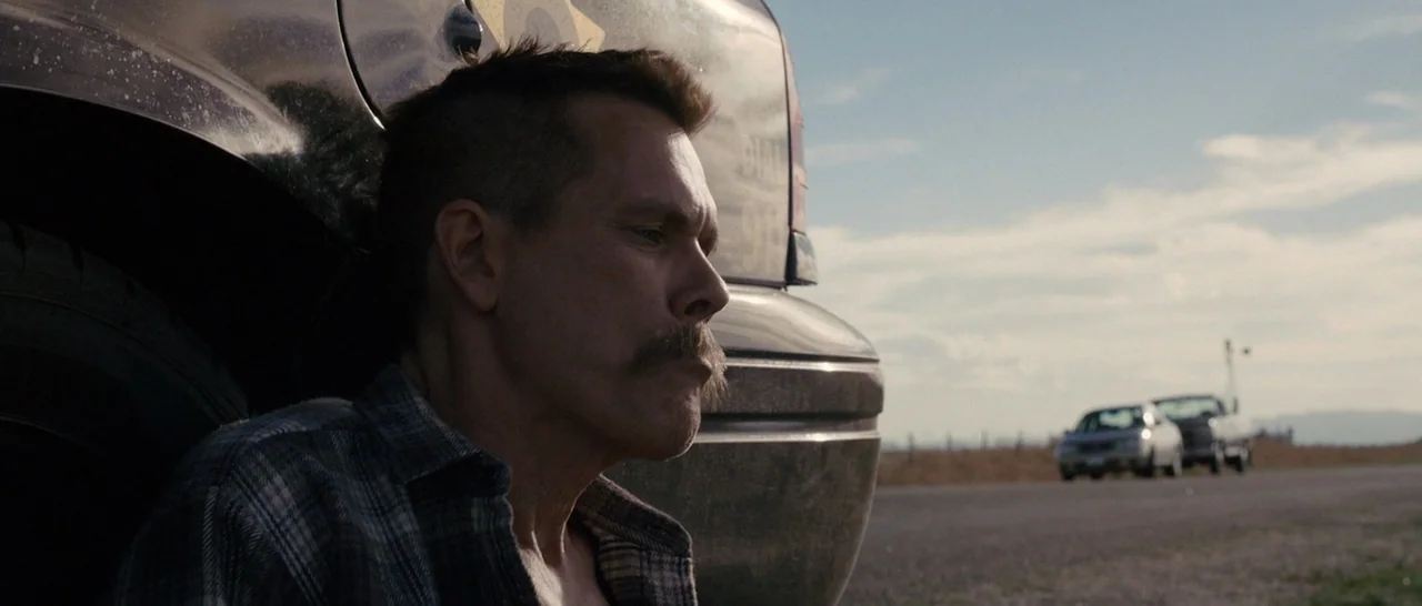 Cop Car (2015) movie still: medium — A man sits on the shoulder of a desert highway against a dark vehicle. Two cars are parked in the distance…