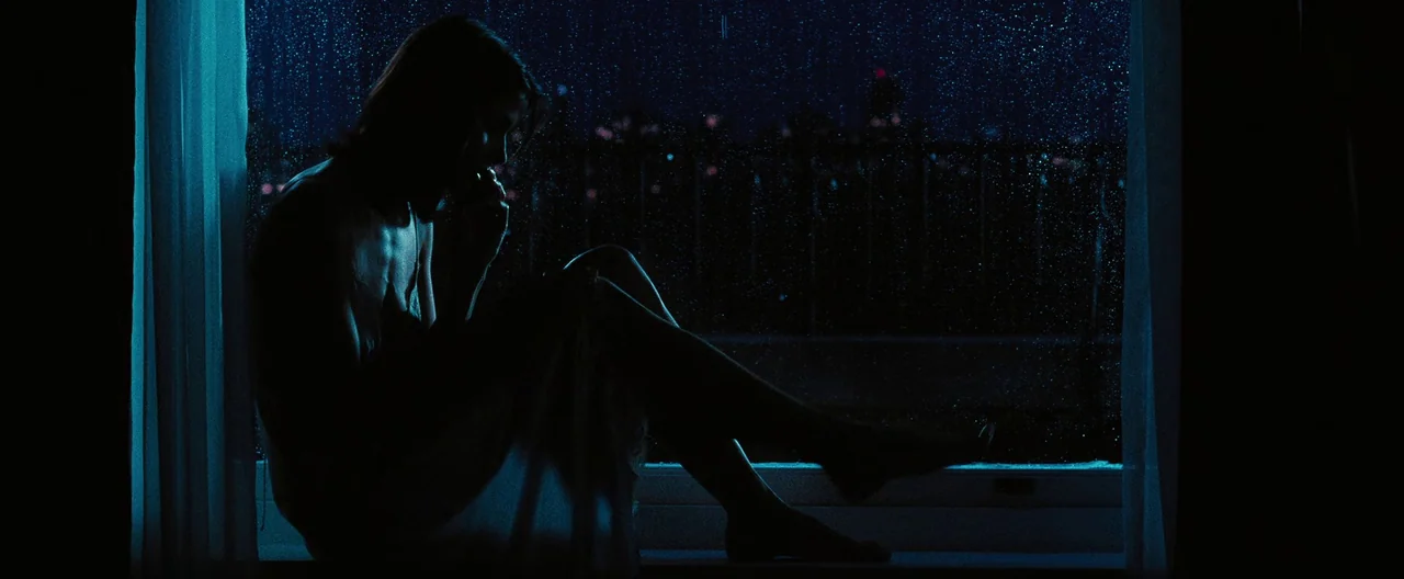 Lord of War (2005) movie still: fullbody — A woman sits on a rain-streaked window sill at night overlooking a dark city. She is silhouetted against…