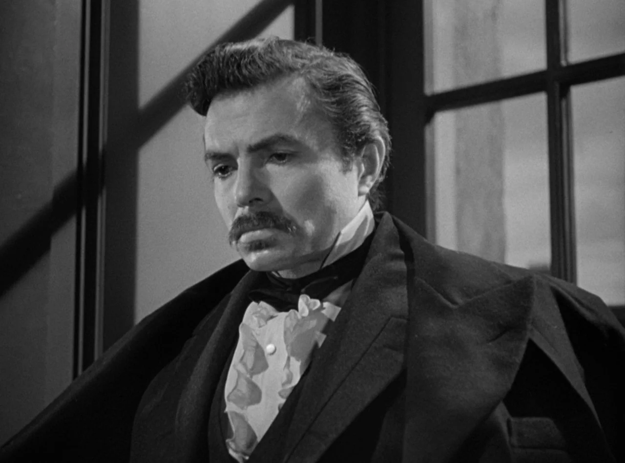 Madame Bovary (1949) movie still: medium — A man in formal attire stands near a window frame indoors at night. The man gazes downward while wearing…