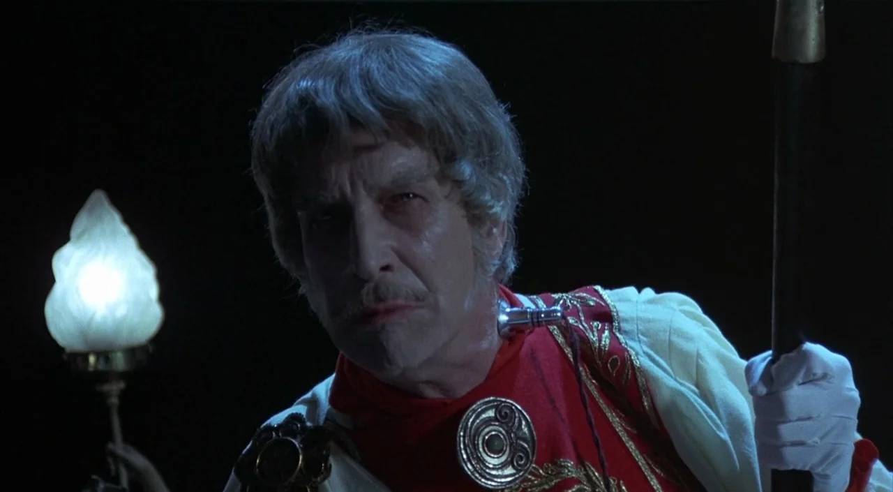 Dr. Phibes Rises Again (1972) movie still: closeup — A man in ornate period clothing stands against a pitch-black background in darkness. He holds a…