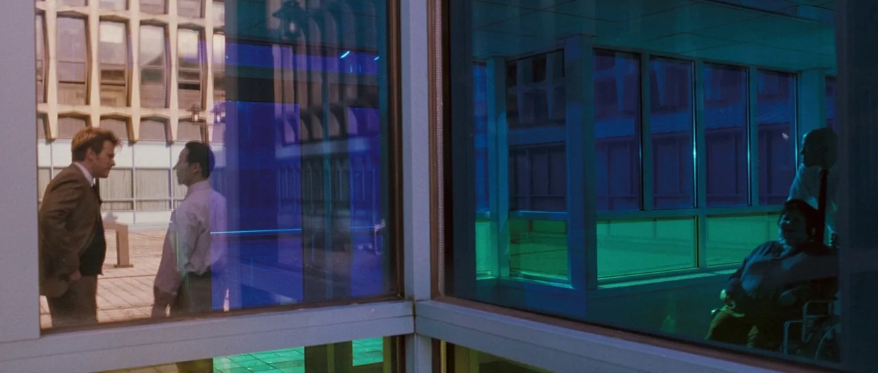 Stay (2005) movie still: wide — A modern building exterior seen through layered glass windows with blue and green reflections. Two men stand on a…