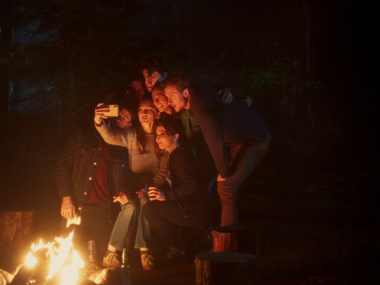 In A Violent Nature (2024) movie still: medium — A group of young adults huddles together at night in a dark forest setting. They lean in close…