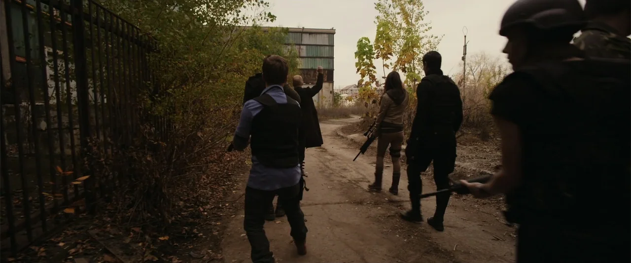 The Condemned 2 (2015) movie still: fullbody — An overgrown dirt path runs between a high iron fence and an industrial site. Several individuals in…