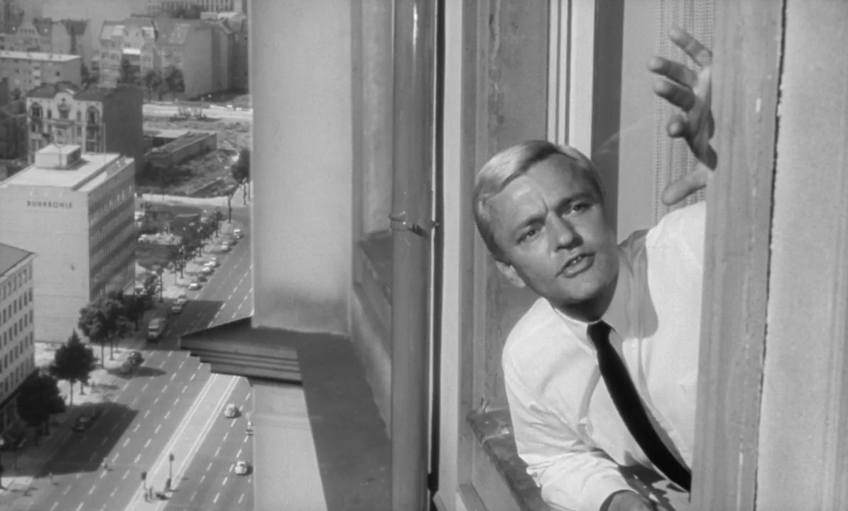 The 1,000 Eyes of Dr. Mabuse (1960) movie still: medium — A man looks out from a high-rise building over a sparse urban street. Cars are parked and…