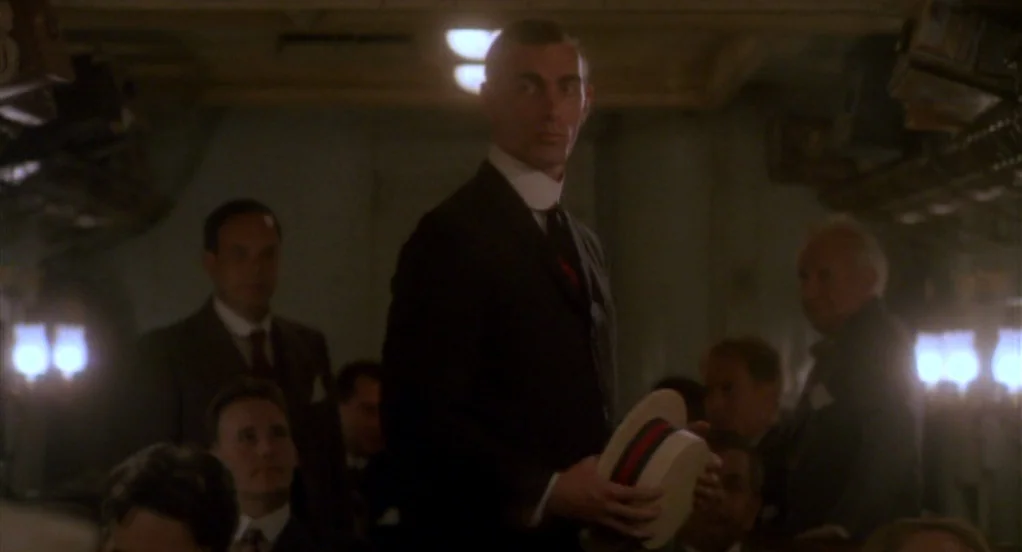 Eight Men Out (1988) movie still: medium — A dimly lit passenger cabin with rows of seated people in formal attire. The man stands in the central…