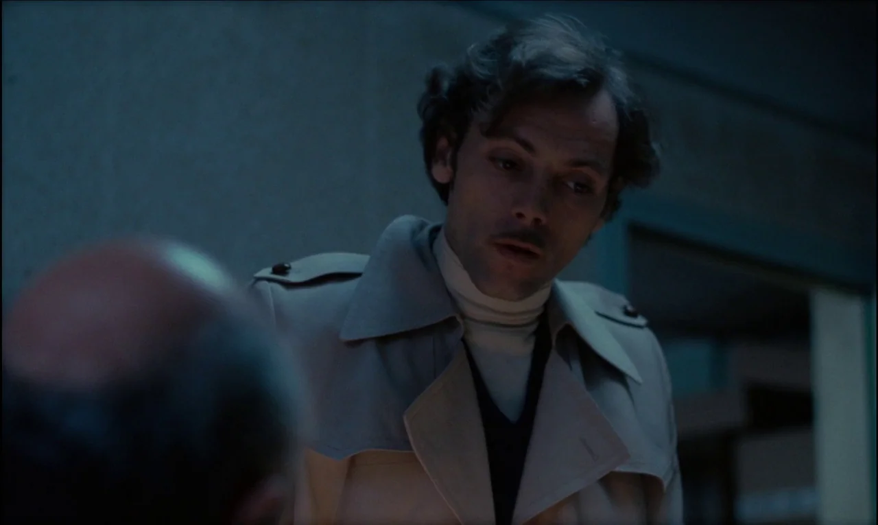 Serie Noire (1979) movie still: medium — A dark office environment features muted walls and dim, low-key lighting. A man in a trench coat leans…