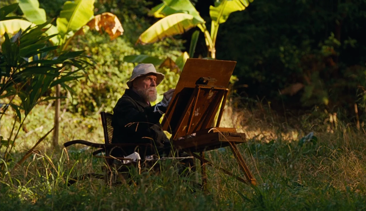 Renoir (2012) movie still: medium — An elderly man paints on a wooden easel positioned in a grassy, lush clearing. Banana leaves and dense foliage…