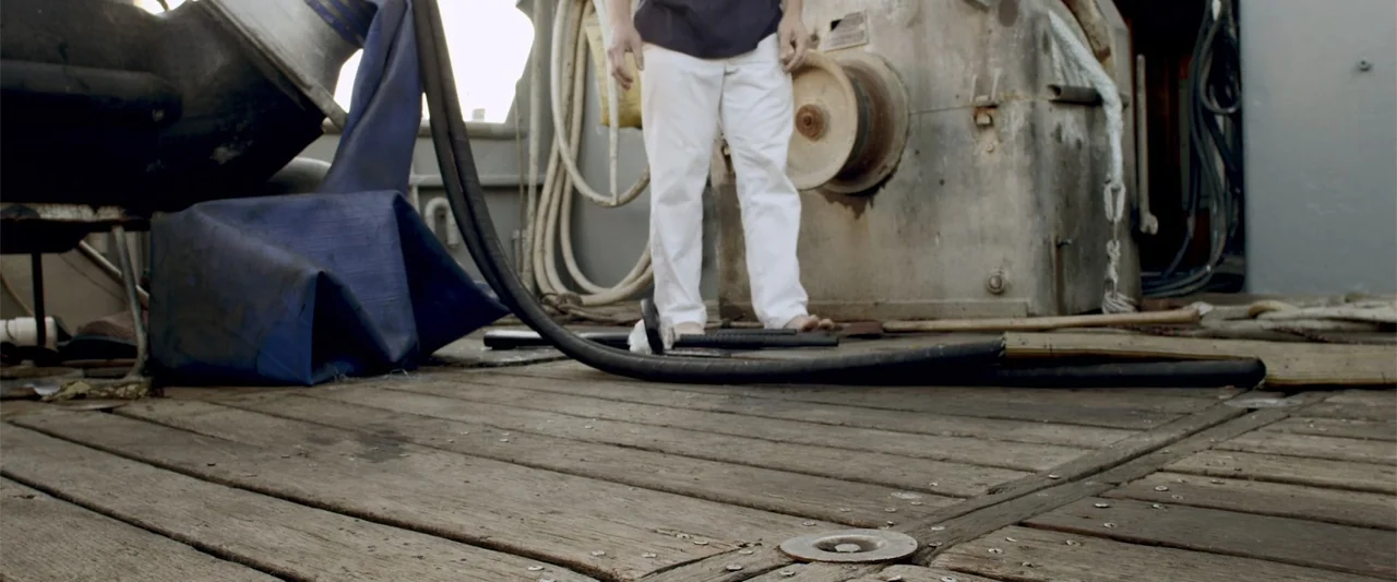 Mermaid Down (2019) movie still: fullbody — The deck of a boat is constructed from weathered wood planks with mechanical metal winches. A barefoot…