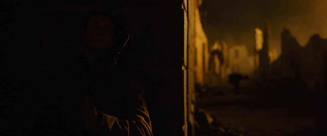 1917 movie still: medium — A soldier stands against a dark corner in a destroyed, fire-lit urban landscape. The ruins of buildings glow intensely…