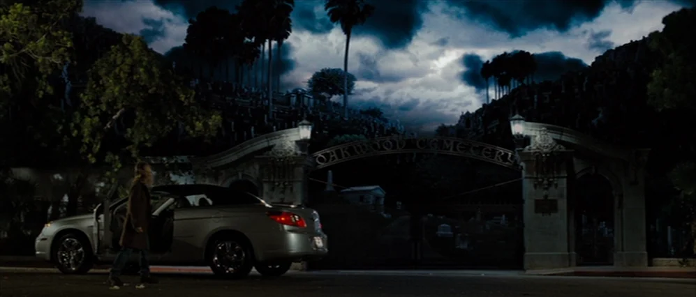 Drag Me to Hell (2009) movie still: wide — A silver convertible sits before a wrought iron and stone cemetery gate at night. A woman stands beside…