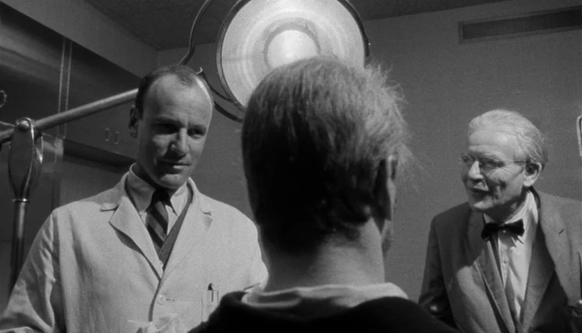 Seconds (1966) movie still: medium — A clinical exam room features a bright overhead lamp and metallic wall fixtures. Two men speak to a person…