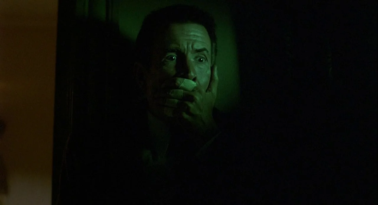 Psycho III (1986) movie still: closeup — A man stands in a dark space lit by a greenish light source. The man holds his hand over his mouth with a…