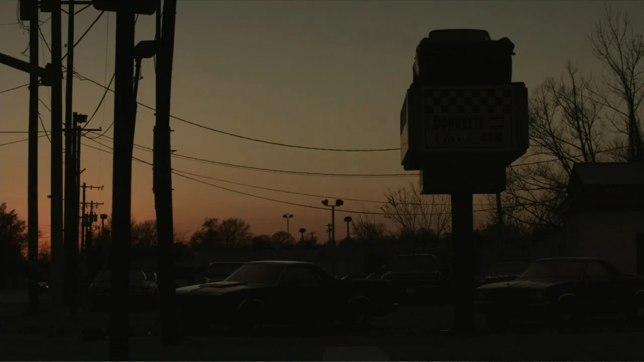 Death of a Child (2017) movie still: establishing — An exterior view of a car lot at dusk with silhouettes of vehicles. Utility poles and a tall…
