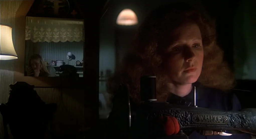 Carrie (1976) movie still: medium — A dimly lit room contains a vintage metal sewing machine in the foreground. A woman with auburn hair uses the…