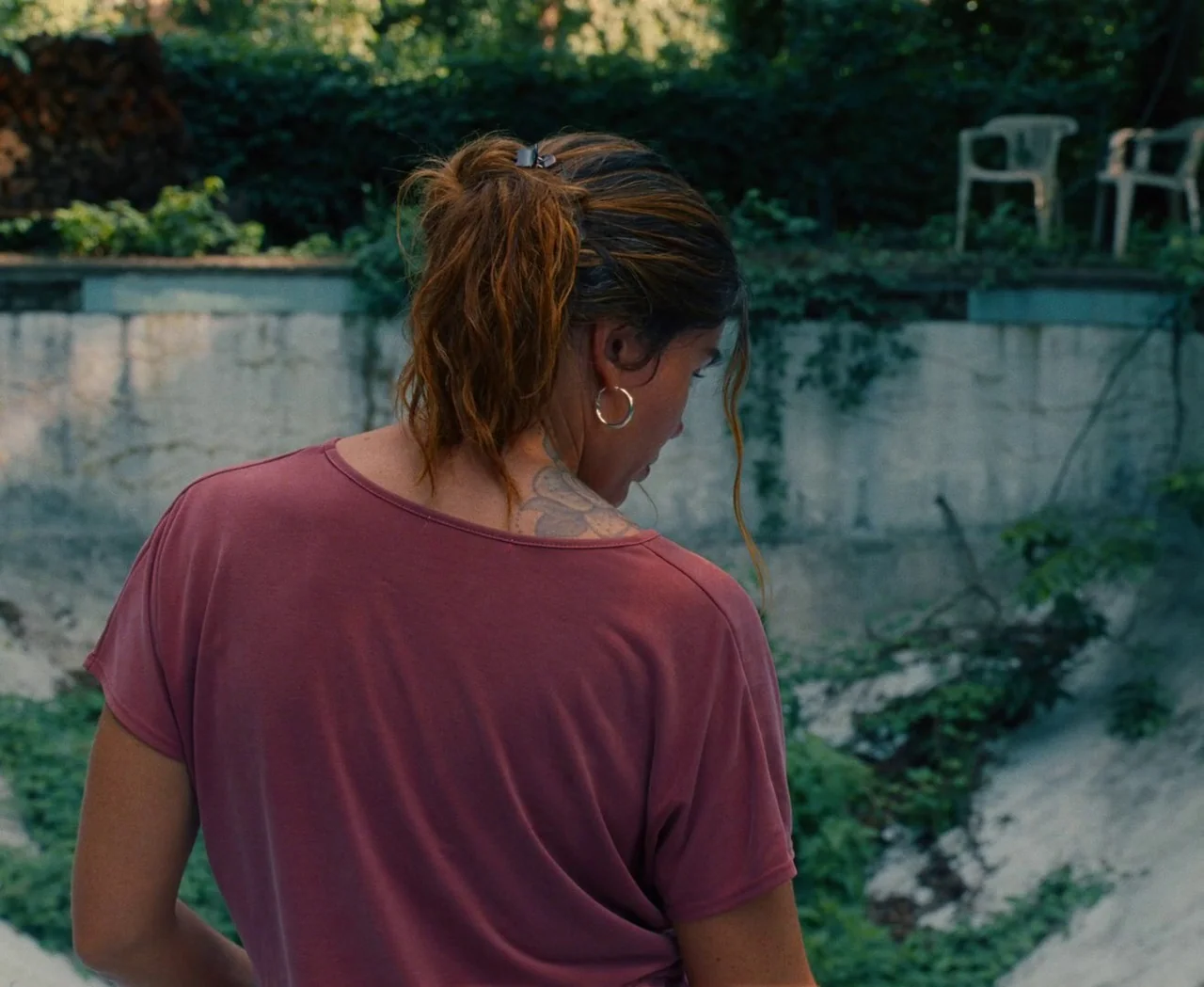 Monica (2022) movie still: medium — An empty concrete swimming pool is overgrown with weeds and shaded by trees. A woman in a pink shirt stands at…