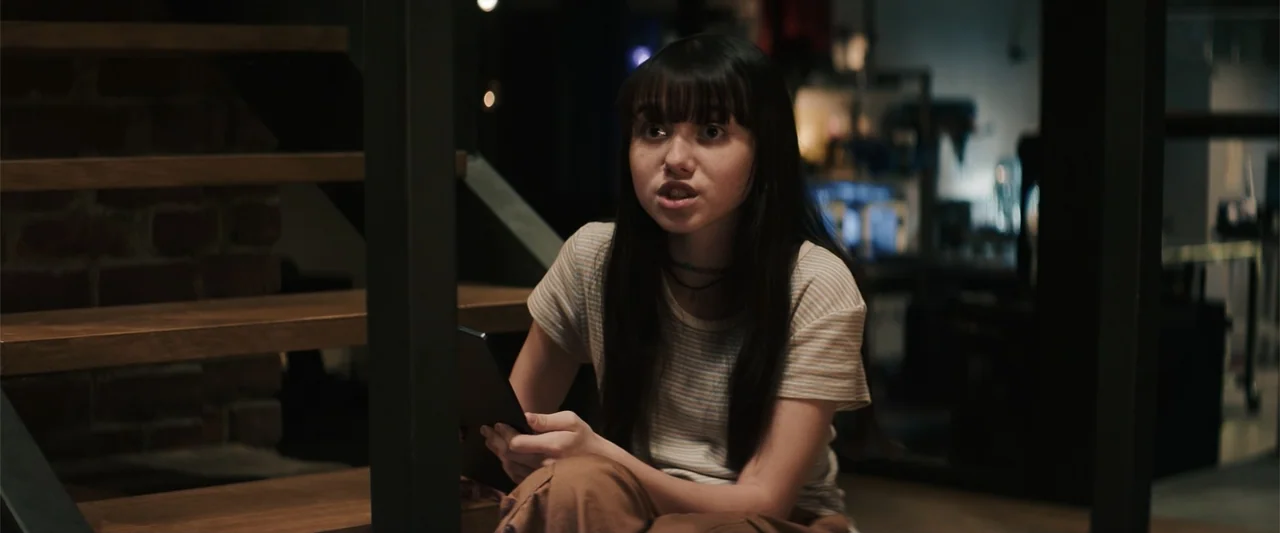 M3GAN 2.0 (2025) movie still: medium — A person sits on a wooden staircase in a dimly lit residential interior. The individual looks forward while…