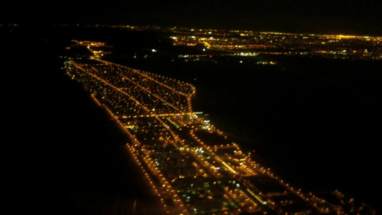 Counting (2015) movie still: establishing — An aerial view of a brightly lit city grid surrounded by dark wilderness. Thousands of streetlights form…