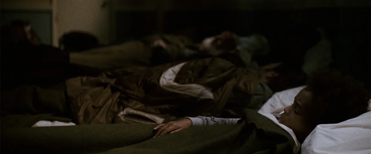 The Pursuit of Happyness (2006) movie still: medium — A dimly lit room contains multiple simple cots occupied by people sleeping. A young child lies…