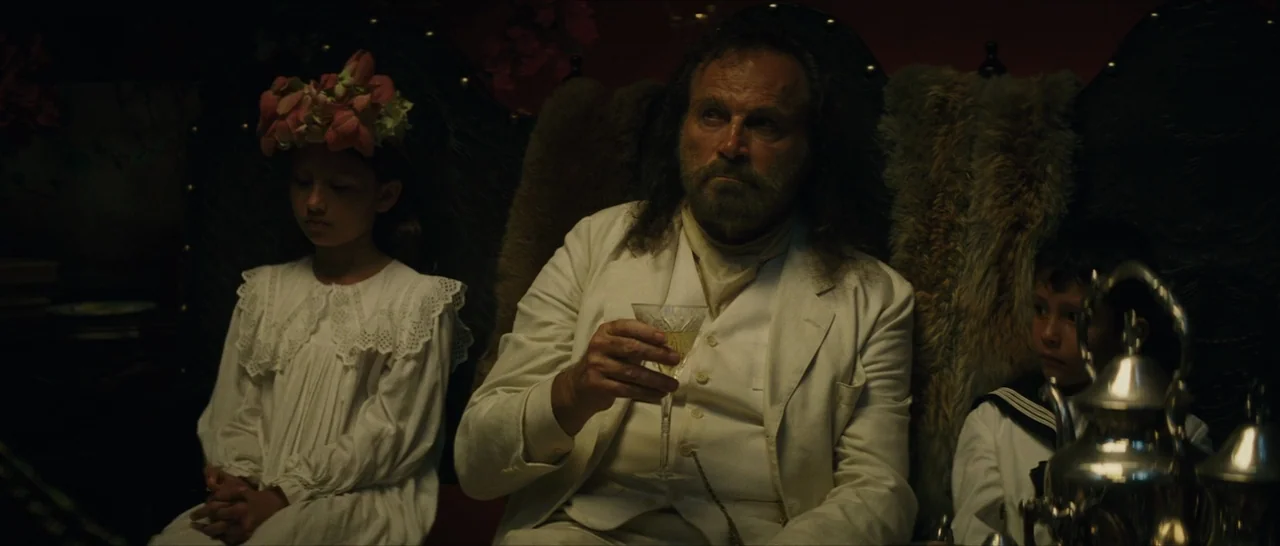 The Lost City of Z (2016) movie still: medium — A dimly lit room features a man and two children sitting on ornate furniture. The man holds a…