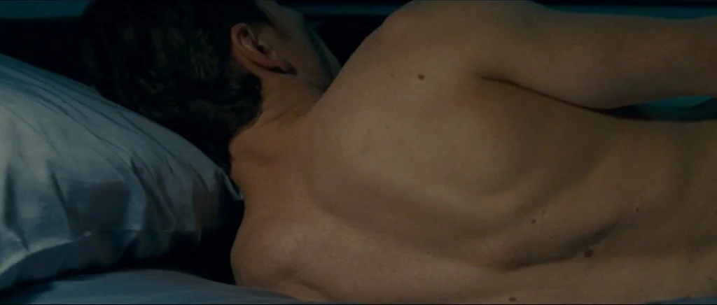 Shame (2011) movie still: closeup — A person rests on a blue bed sheet in a dimly lit room. The subject's bare back is positioned against a soft…