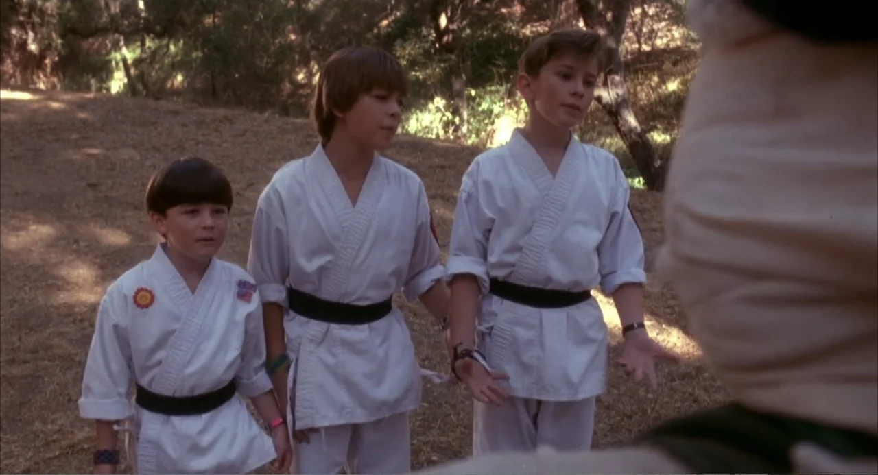 3 Ninjas (1992) movie still: medium — Three young boys in white martial arts uniforms stand in a sunlit woodland area. The boys face an unseen…
