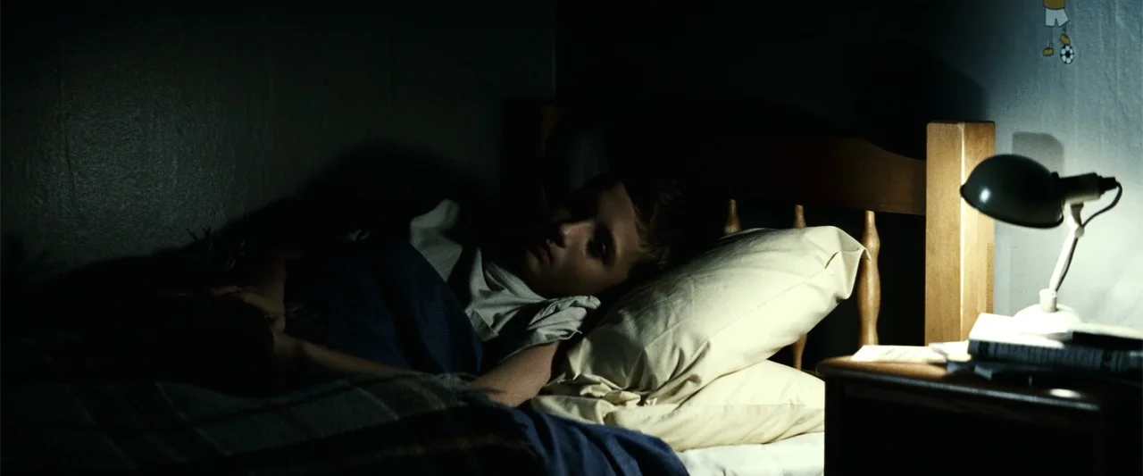 Hereafter (2010) movie still: medium — A dark bedroom at night features a bed frame against a plain wall. A young boy lies under a blue blanket with…