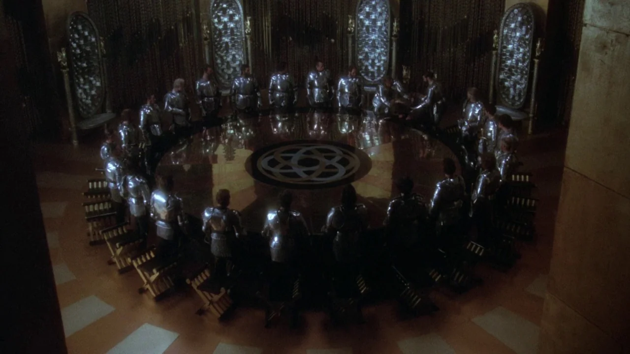 Excalibur (1981) movie still: wide — A high-angle view shows numerous knights in silver plate armor seated around a table. The large table features…