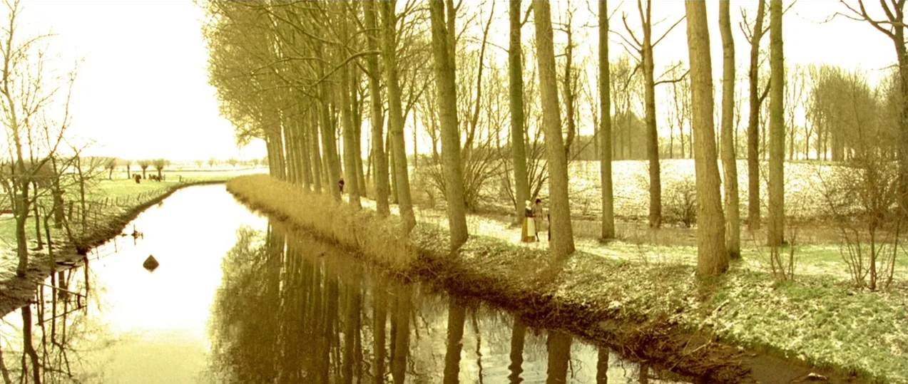 Girl with a Pearl Earring (2003) movie still: wide — A canal flows through a line of bare trees on a grassy field. Two distant figures in period…