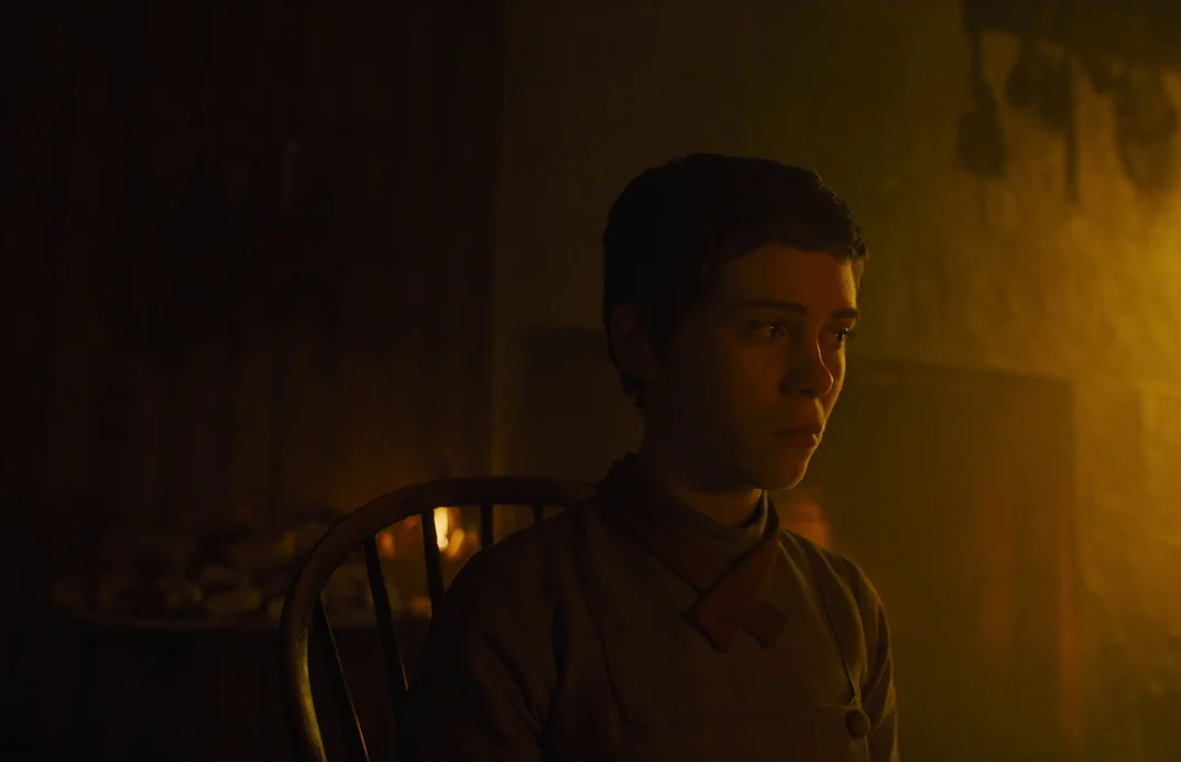 Gretel & Hansel (2020) movie still: medium — A dimly lit room with textured dark walls and a rustic wooden chair. A young woman with short hair sits…