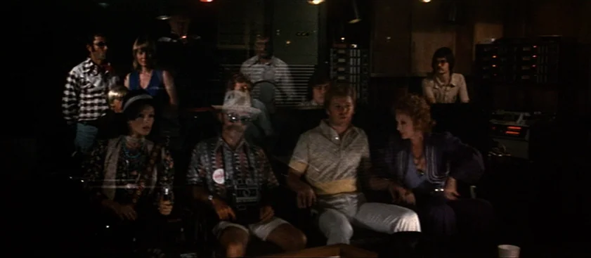 Nashville (1975) movie still: medium — A dimly lit industrial room contains rows of rack-mounted electronic equipment and panels. Several adults sit…
