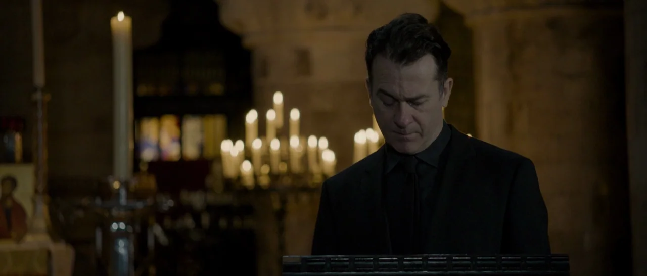 The Face of an Angel (2014) movie still: medium — A man stands inside a dimly lit stone building with many candles glowing behind. He is looking…