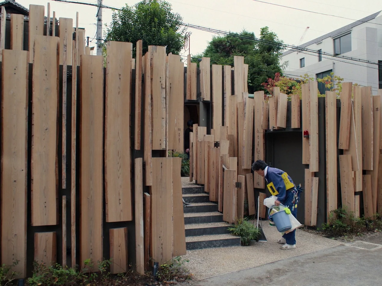 Perfect Days (2023) movie still: wide — An exterior wall clad in vertical raw timber planks hides a small staircase entrance. A man in a work…