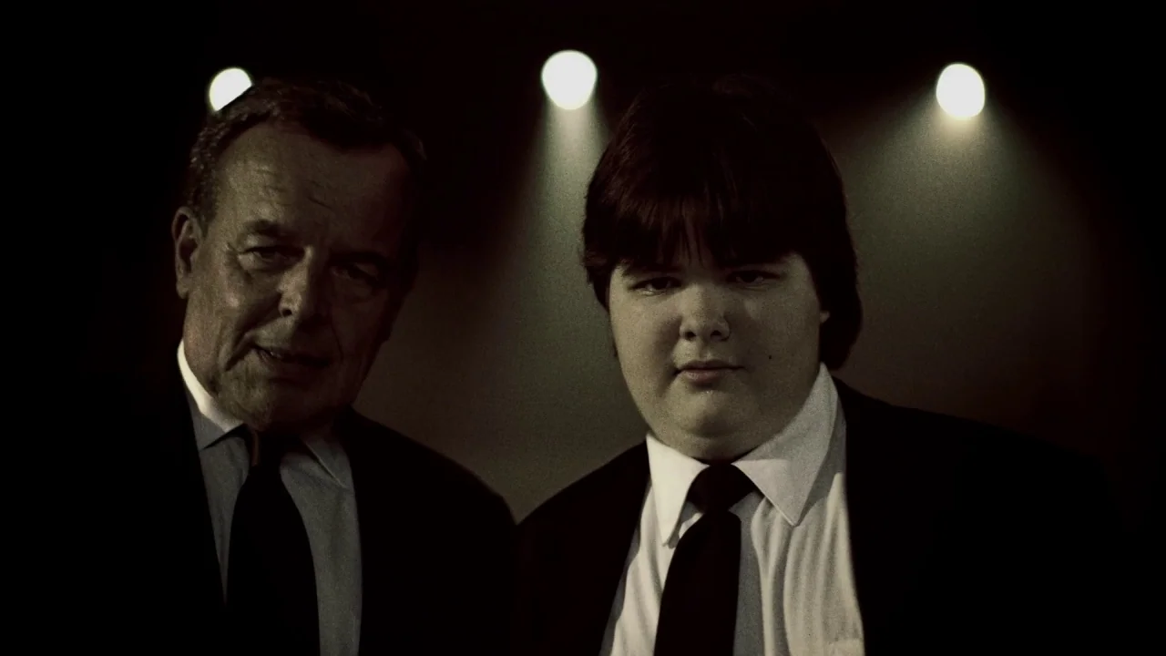 Suburban Gothic (2014) movie still: medium — Two men stand against a pitch-black background with three bright lights behind them. The older man…