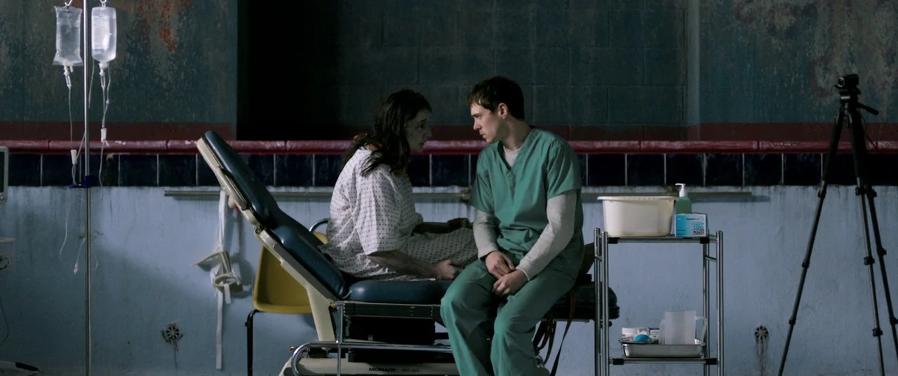The Cured (2017) movie still: medium — A clinical space with tiled walls features an exam table under fluorescent lighting. A man in medical scrubs…