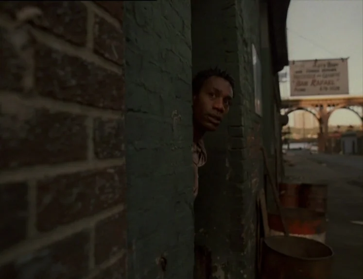 The Brother From Another Planet (1984) movie still: medium — A dark green brick wall creates a frame within the frame on the street. A man peeks…