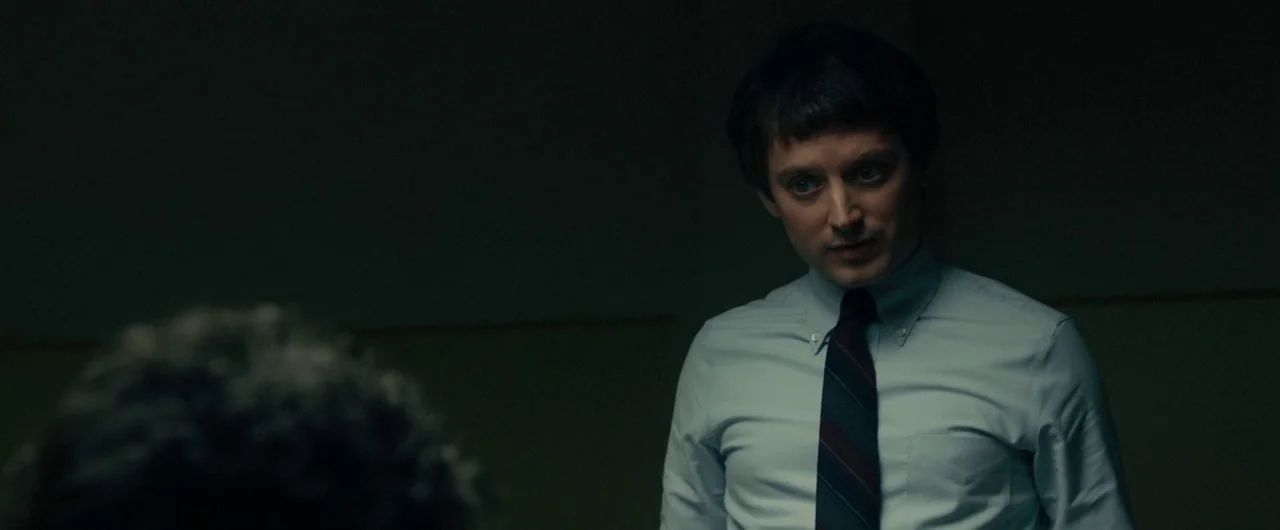 No Man of God (2021) movie still: medium — A dimly lit room with plain dark green walls and minimal furniture. A man in a collared shirt faces an…