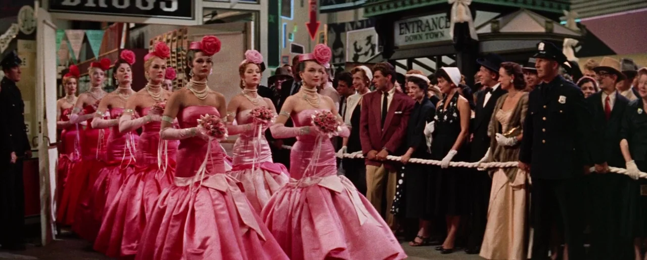 Guys and Dolls (1955) movie still: wide — A parade or public event takes place in a decorated city street setting. A group of women in pink dresses…