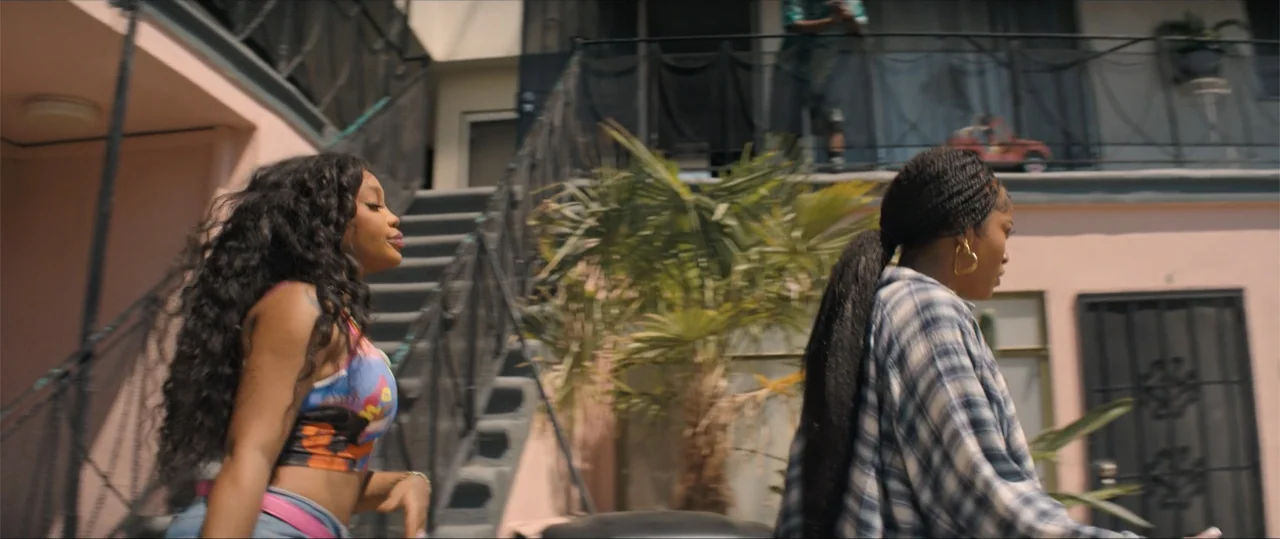 One of Them Days (2025) movie still: medium — A pink exterior apartment building with metal stairs and a central palm tree. Two women walk in the…